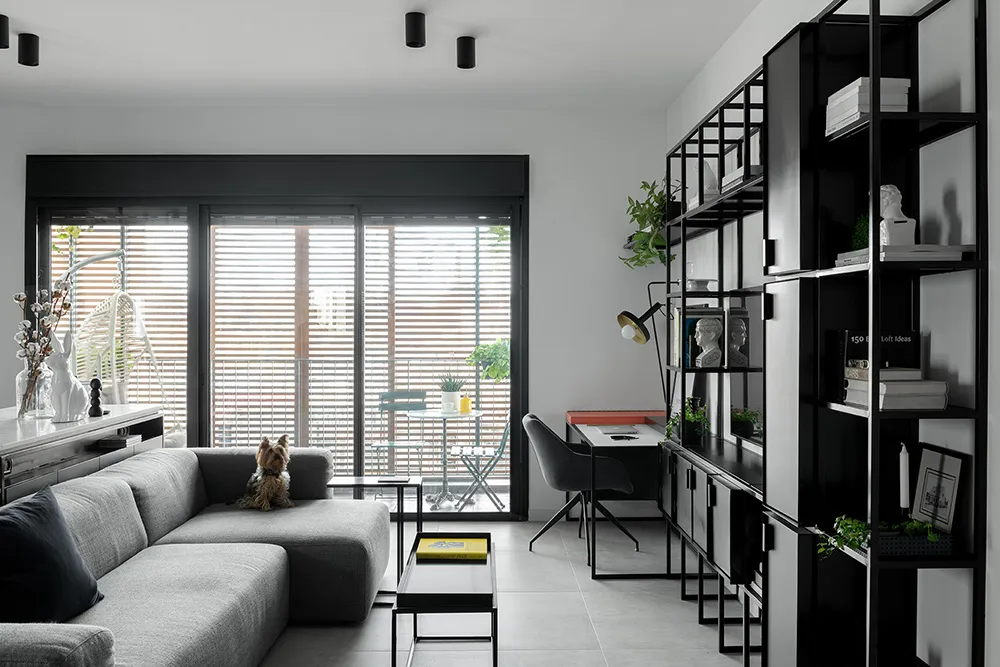 A modern minimalist living room featuring a grey sectional sofa, a glass coffee table, and a black and white shelving unit wi