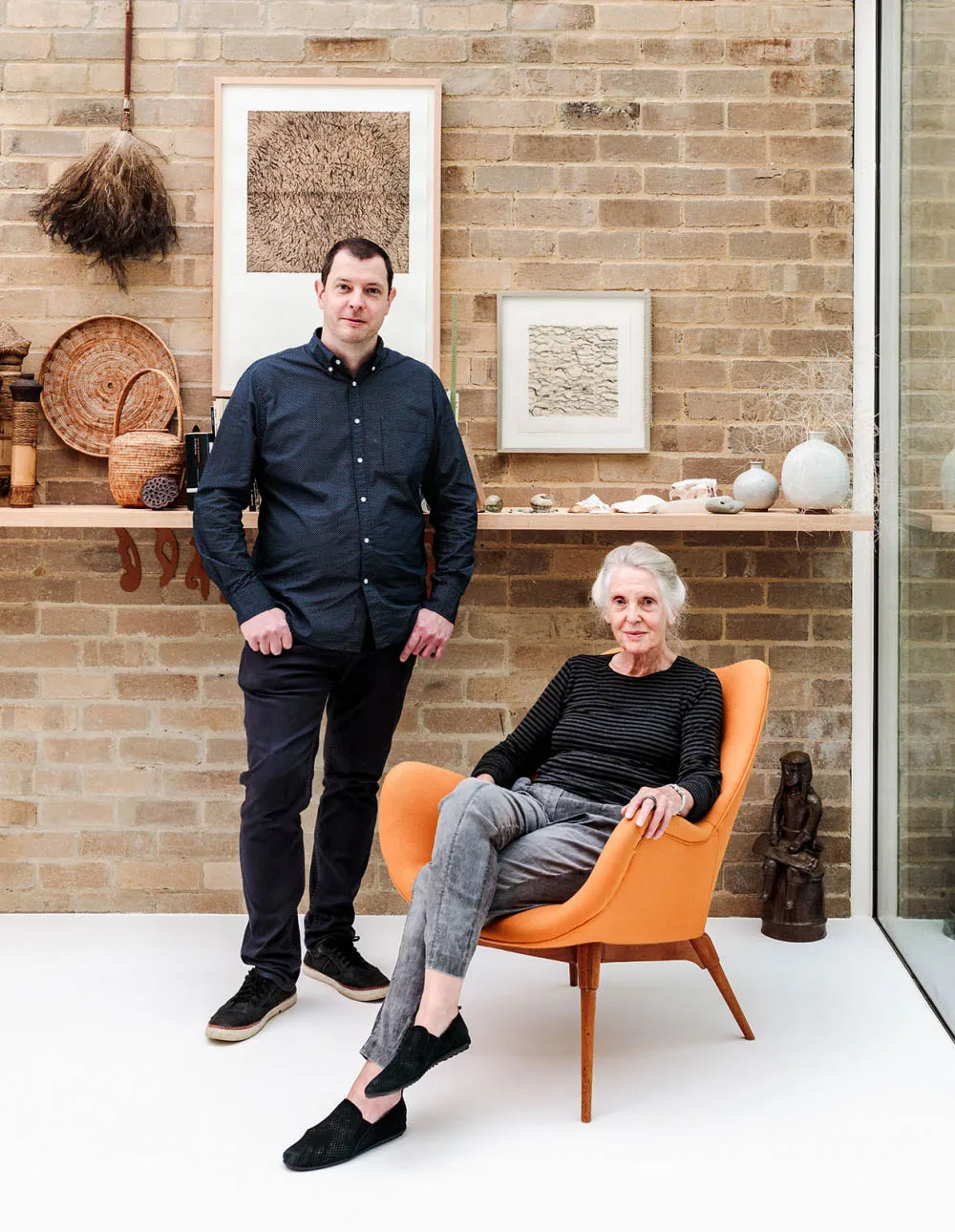 A man and a woman seated in a modern home office with a white floor, brick walls, and a glass partition.
