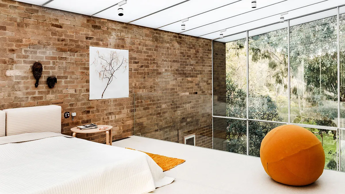 A minimalist bedroom with walls made of brick and concrete, featuring a large window and an orange pouf.