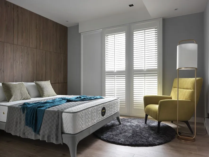 A modern bedroom with wooden accents, featuring a grey bed and yellow armchair, illuminated by natural light through white sh