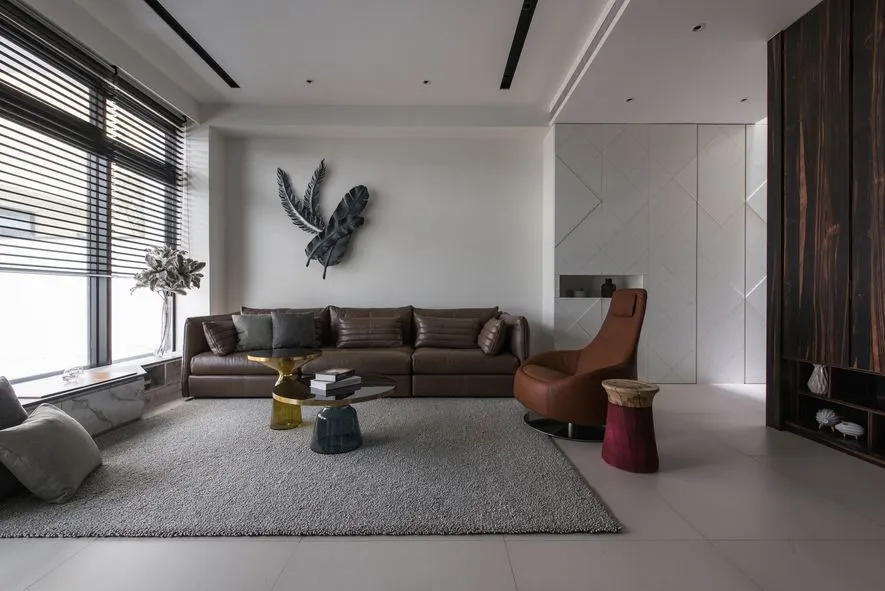A modern living room featuring a brown leather sofa, wooden armchair, glass coffee table, and a decorative wall hanging.
