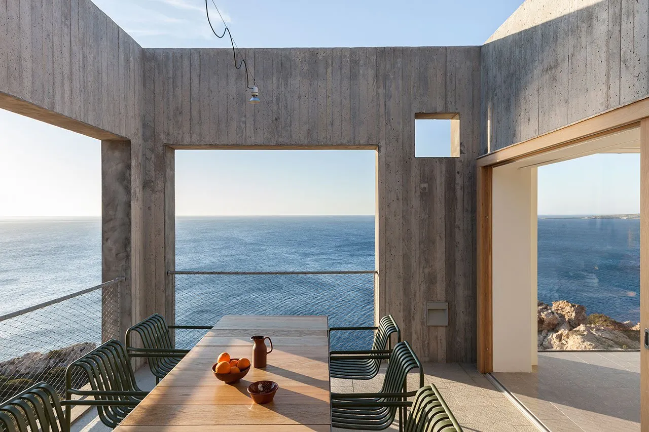 Contemporary outdoor dining area with concrete walls, glass windows overlooking the sea.