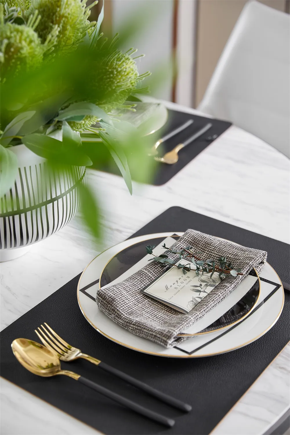 Modern dining table set with glass plates, metal cutlery, greenery centerpiece, and place mats.