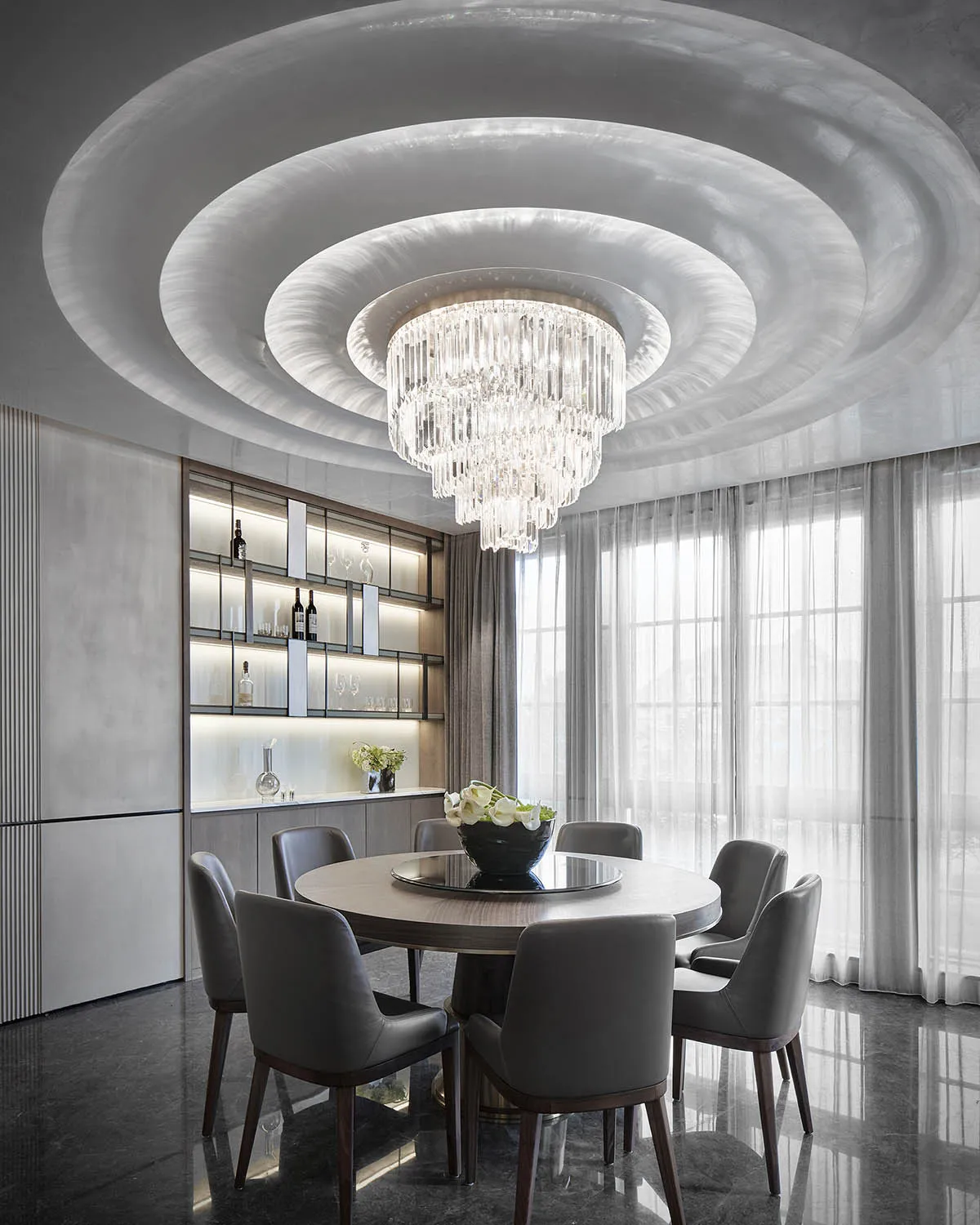 A modern dining room with mixed materials, including a round table, gray chairs, and a crystal chandelier.