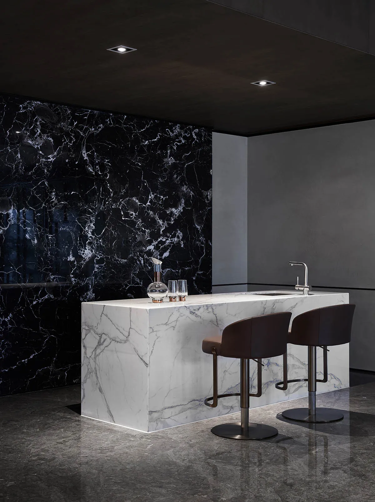 Modern kitchen with white marble countertop and black marble backsplash, featuring a single water faucet and two bar stools.