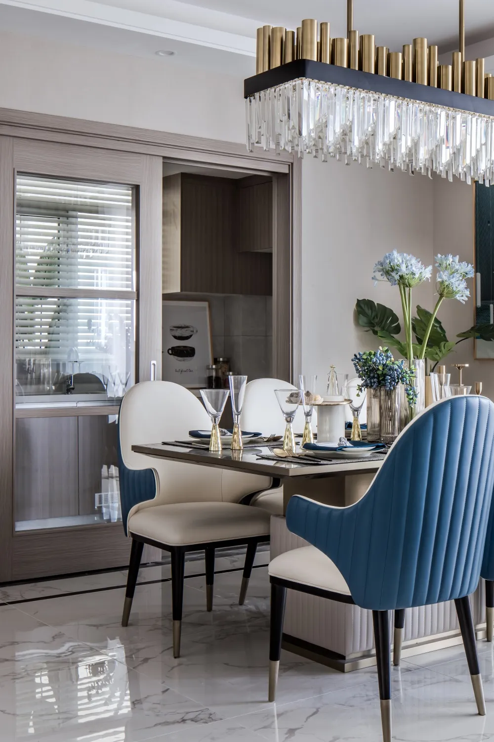 Modern dining room featuring a white marble floor, blue leather chairs, and a gold crystal chandelier.
