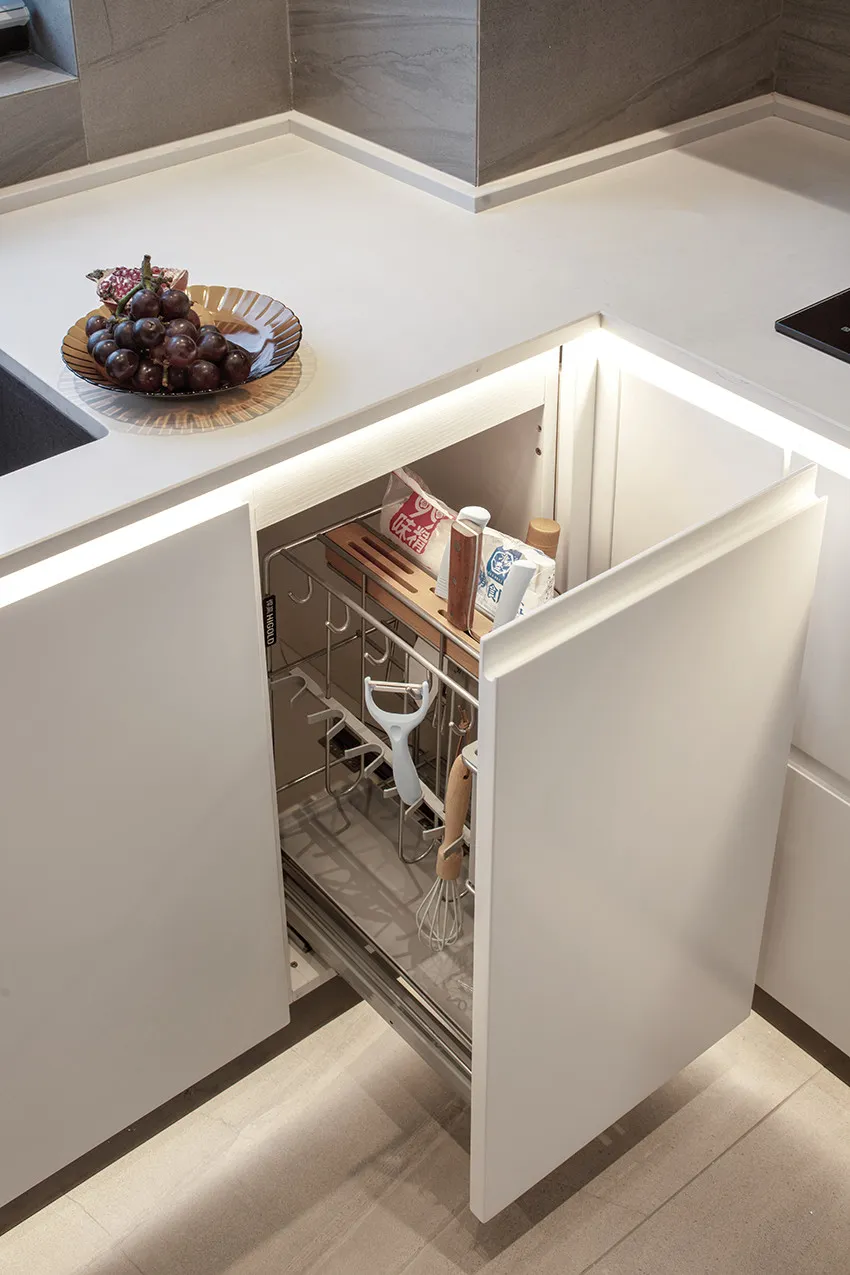 A modern, minimalist kitchen with white cabinetry, featuring an organized drawer and a countertop with grapes and a bowl.