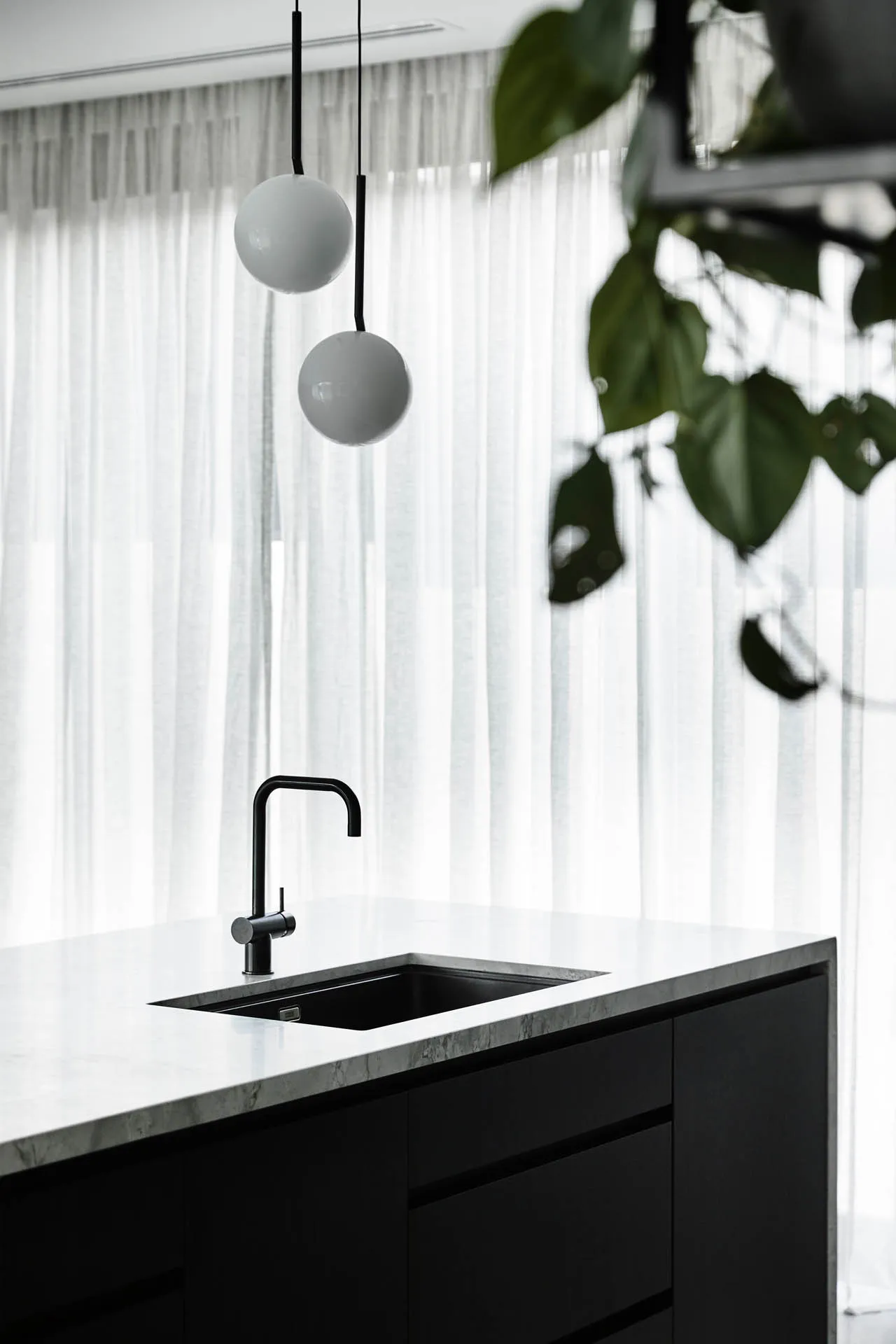 Modern kitchen with black and white marble countertops, black sink, and hanging white globe lights. White sheer curtains and