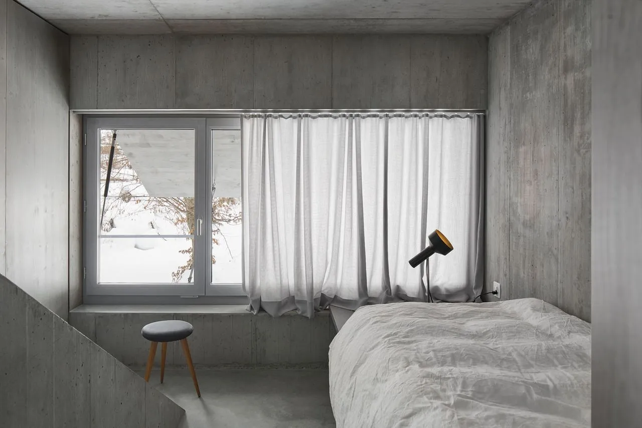 A minimalist bedroom with concrete walls, a white bed, and a white curtain at the window.