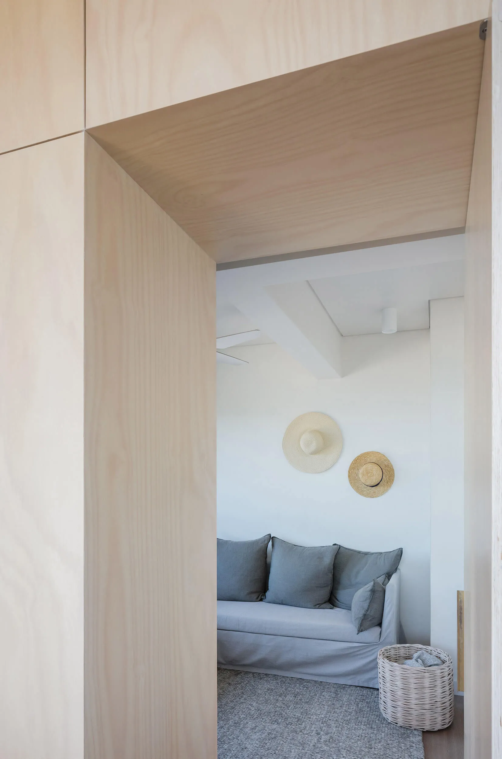 A minimalist living room with a white sofa, grey cushions, and a light wooden wall and floor.