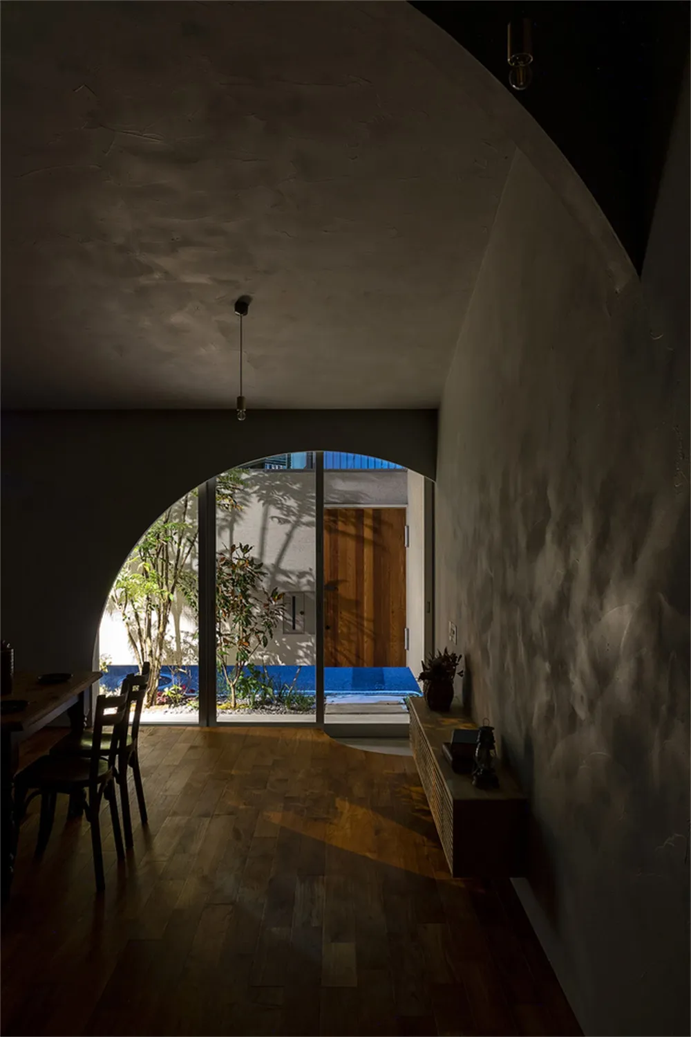 A spacious, modern interior with concrete walls and wooden flooring, featuring a dining area and a view through an archway to