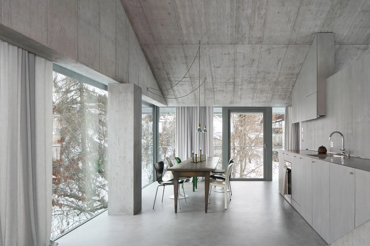 Modern kitchen with concrete walls and floor, large windows showing snowy landscape, and minimalist furniture.