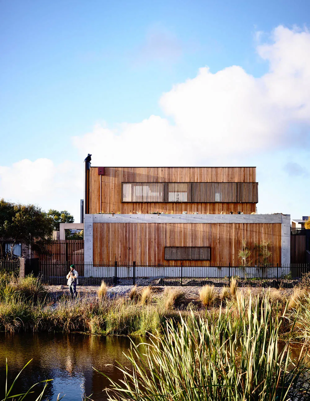 A modern residential home with wooden exterior and large windows, located near a small pond with tall grasses in the foregrou