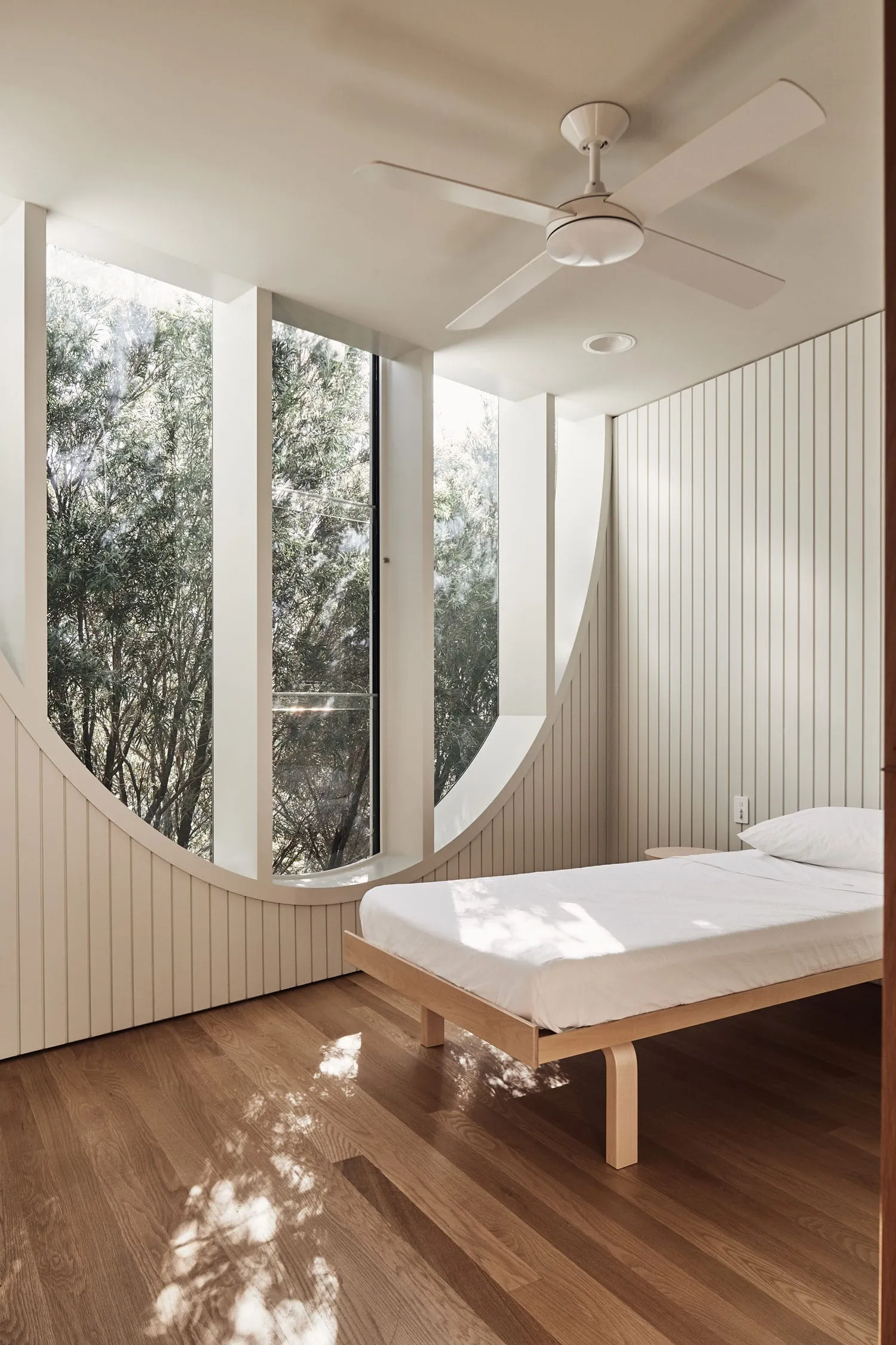 A minimalist bedroom featuring a wooden platform bed in front of a large curved window overlooking trees.