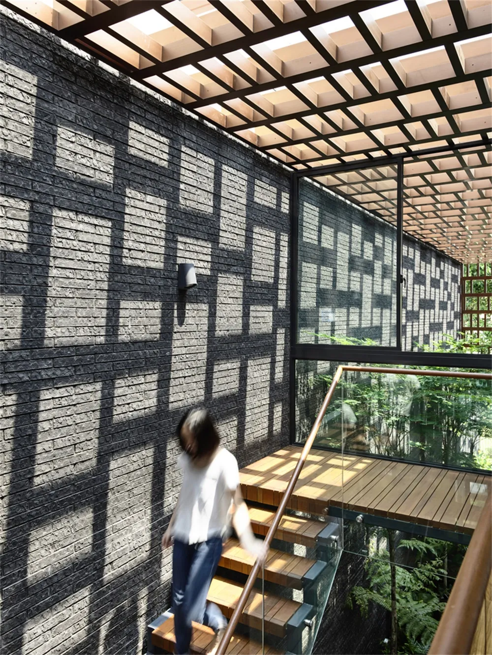 A modern staircase with glass railings and wooden treads, featuring a black brick wall with geometric patterns created by sun