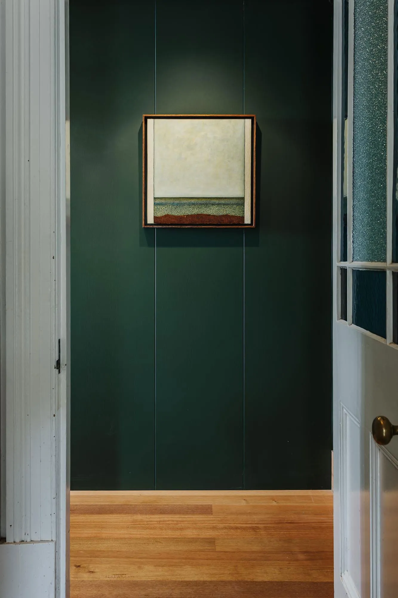 A minimalist interior featuring a framed painting on a dark green paneled wall, with a wooden floor.