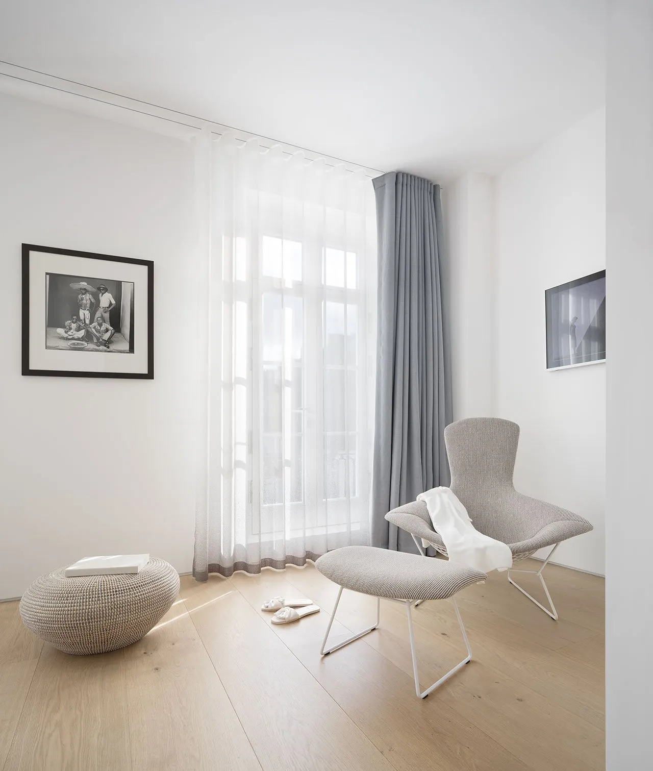A minimalist living room featuring a grey upholstered chair, a white ottoman, and a glass window with sheer curtains.