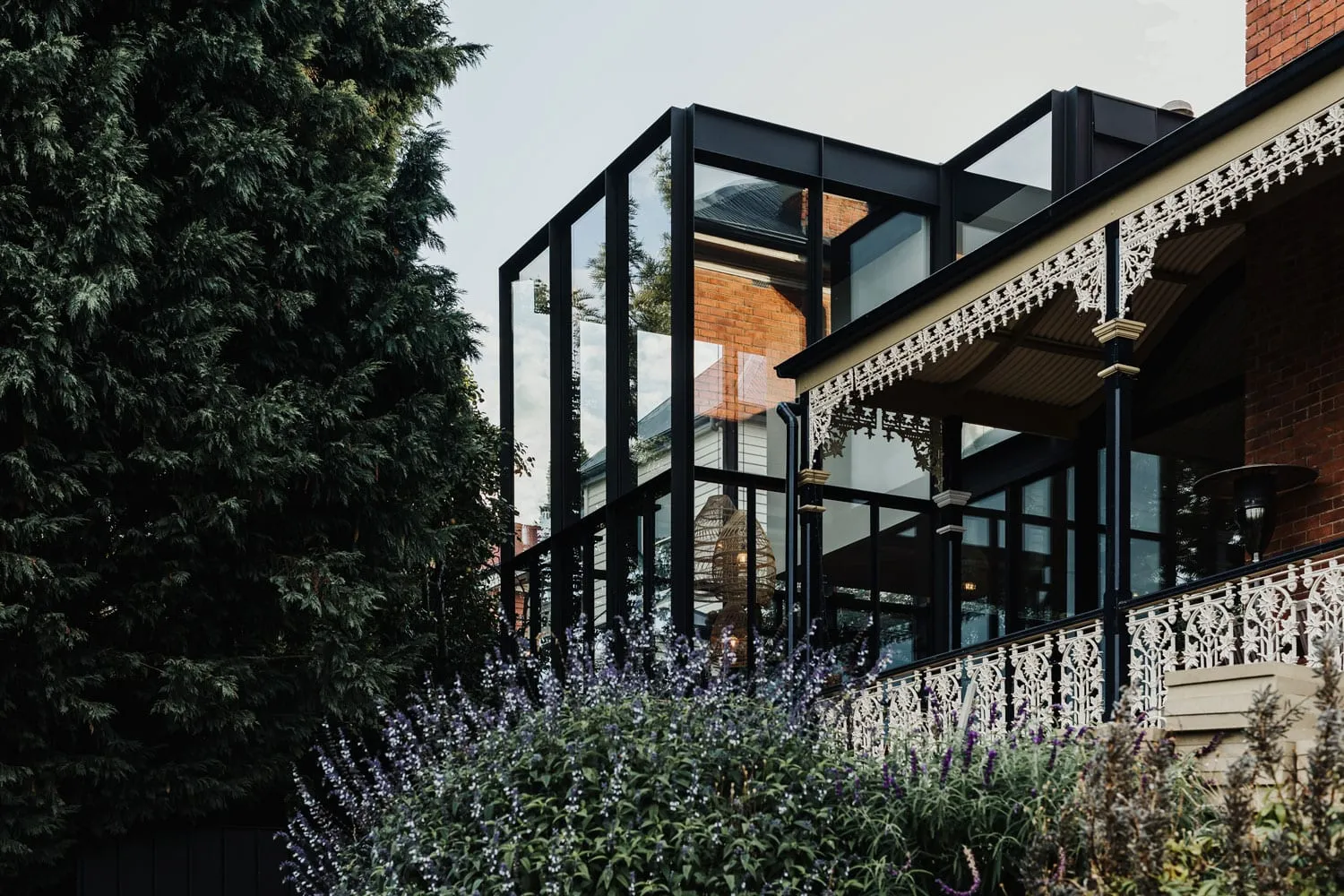A modern house with large glass windows and a brick facade surrounded by lush greenery and purple flowers.