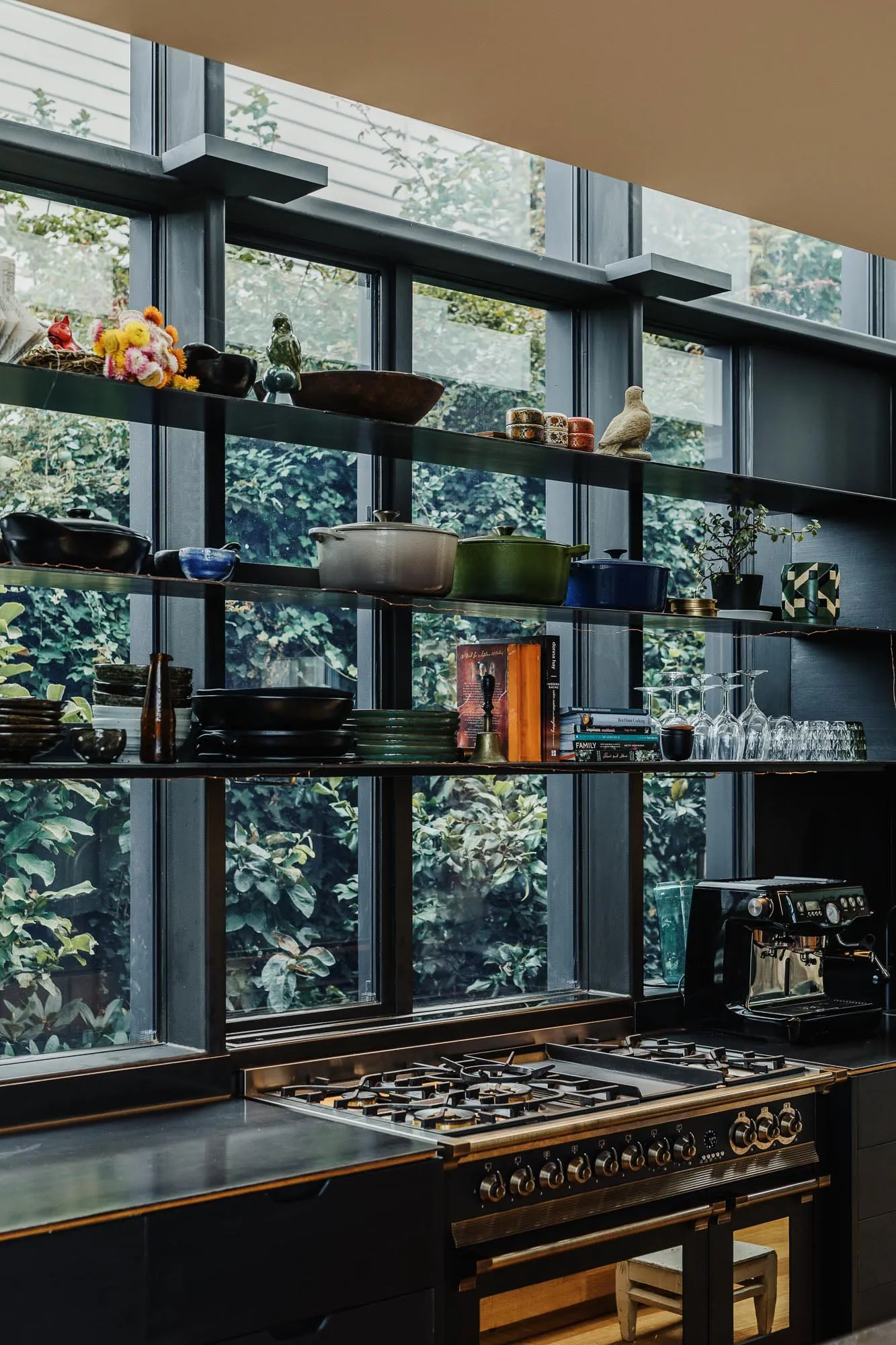 A modern kitchen with metal and glass finishes, featuring a stove, open shelves, and a view of greenery through large windows