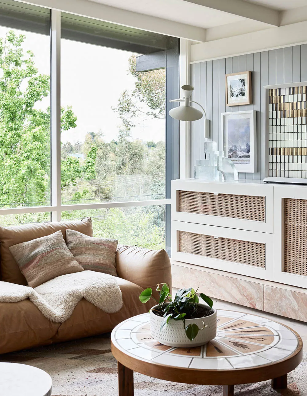 A modern living room with a large window, a tan leather sofa, a round wooden coffee table with a marble top, and a white cabi