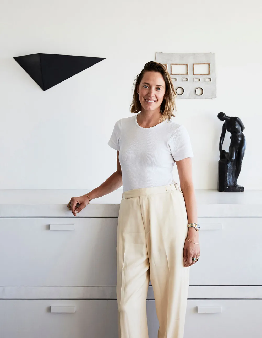 A woman dressed in white standing in front of a white counter with minimalist sculptures and artwork.