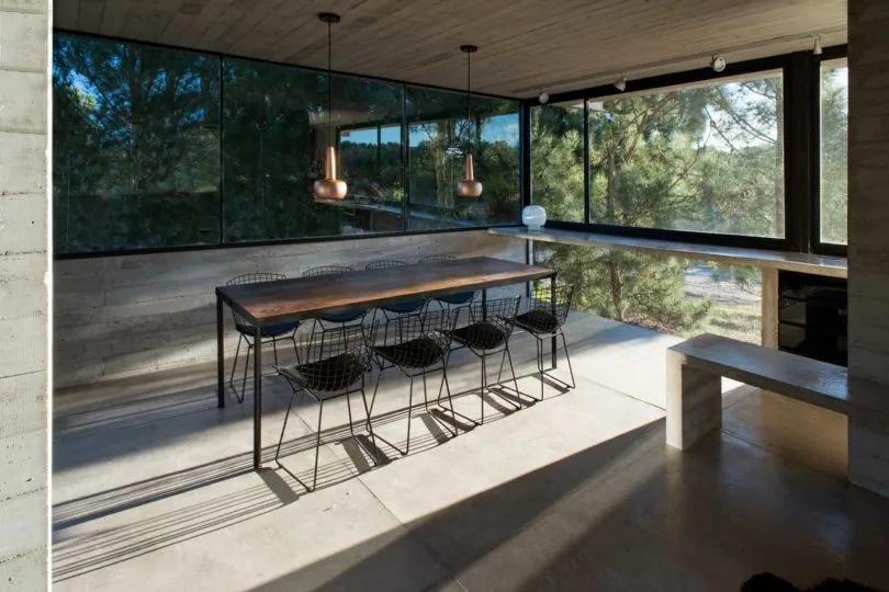 Contemporary dining room with concrete flooring, floor-to-ceiling glass windows, metal chairs, and wooden dining table.
