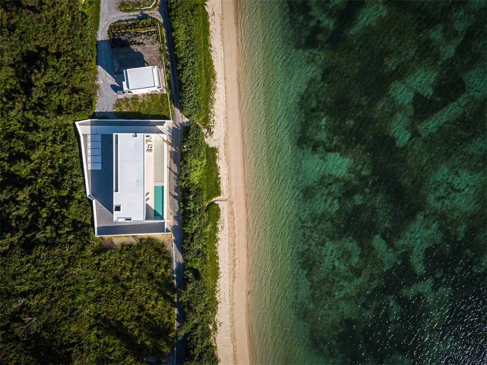 A minimalist beach house with a clay roof, surrounded by greenery and a sandy beach.
