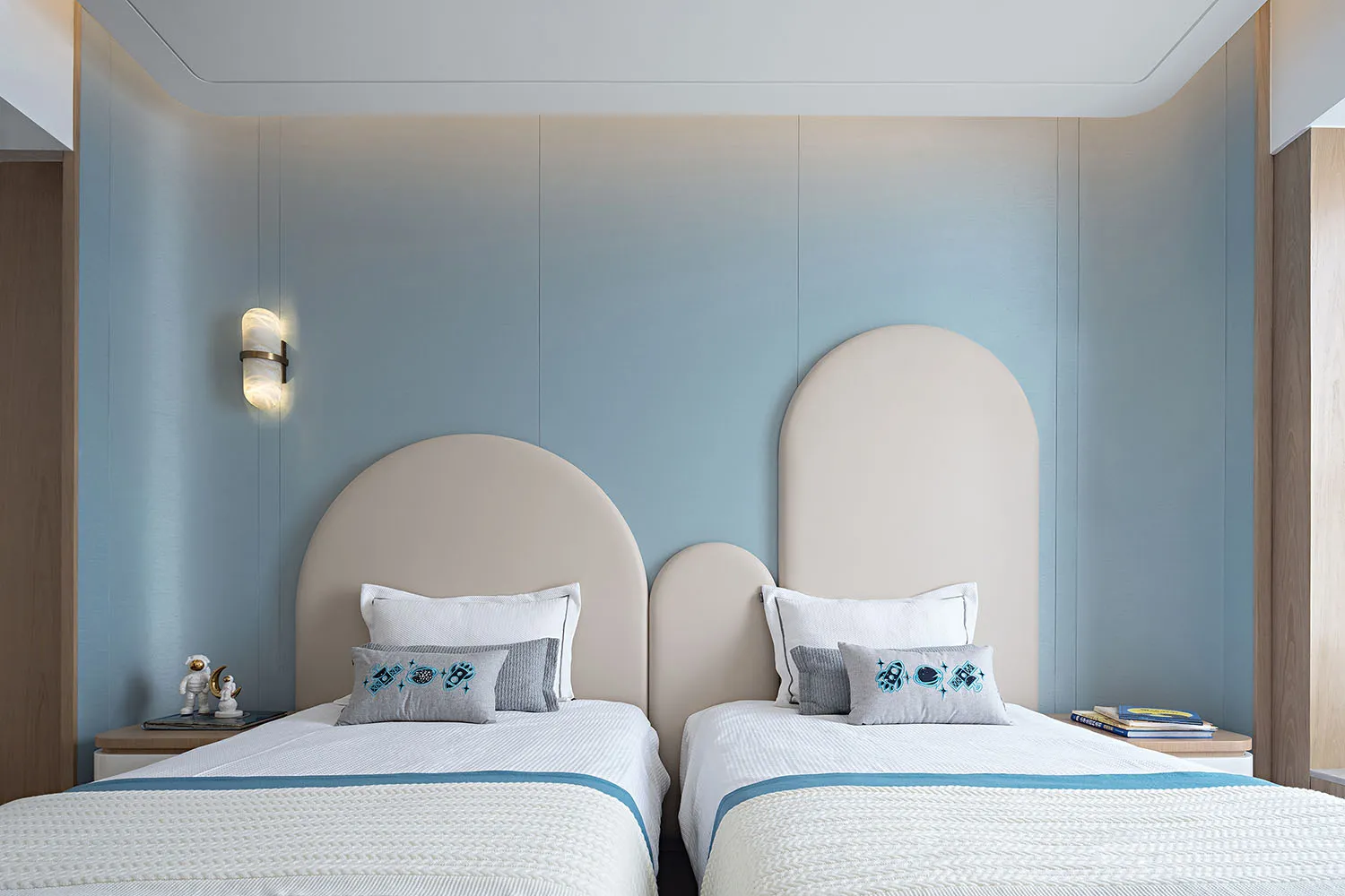 Modern minimalist bedroom with two white beds against blue walls, featuring curved headboards, white bedding, and light blue