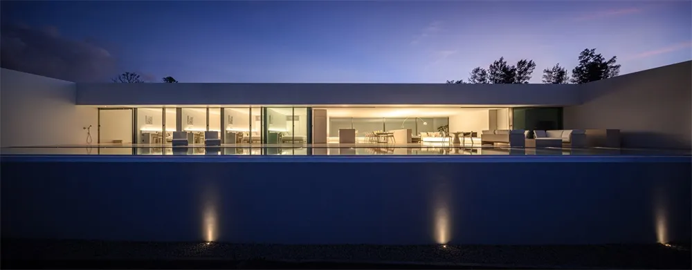 A modern residential exterior with a pool and interior lighting visible through large windows.