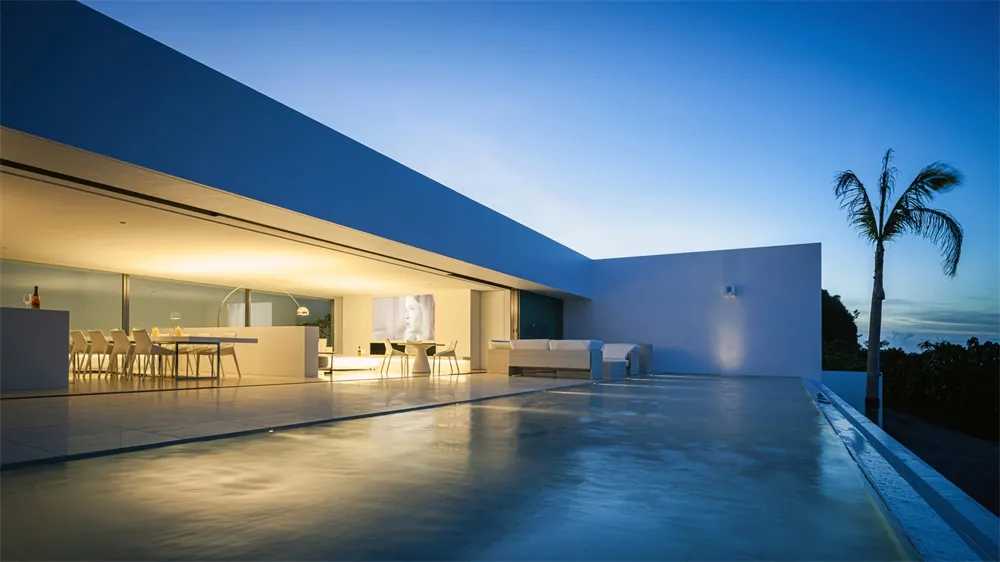 A modern residential patio with a concrete floor and a palm tree, illuminated by lights at dusk.