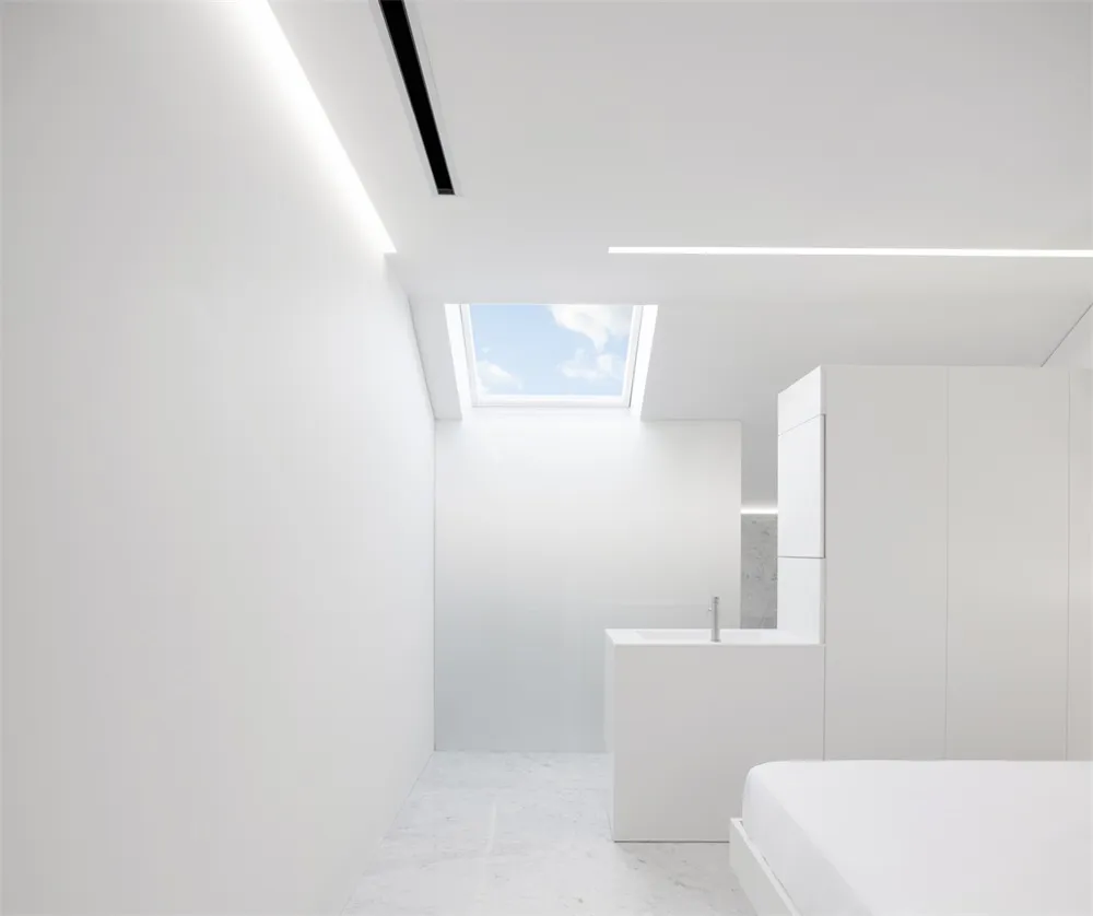 Modern, minimalist bedroom with white walls, ceiling, and floor. Features a large window with natural light.