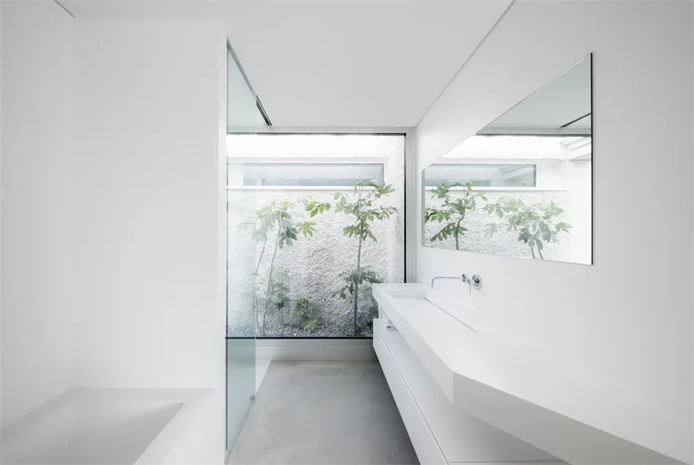 A minimalist bathroom interior with a white vanity, concrete floor, and a window looking into a courtyard with plants.