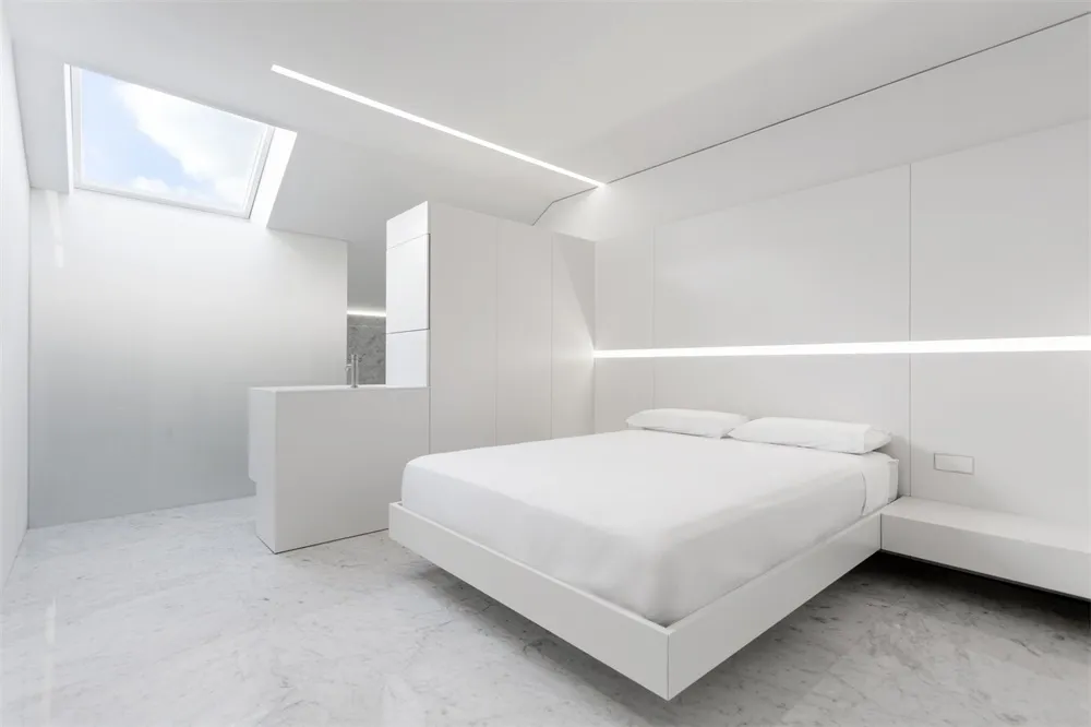 A modern white bedroom with a floating bed, white walls, and a white ceiling light.