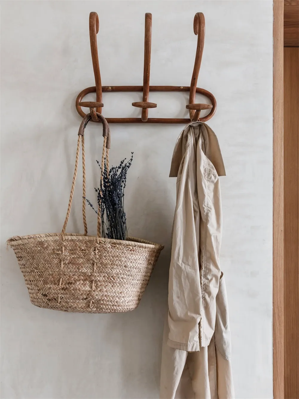 An entryway scene featuring wooden hooks, a woven basket filled with dried flowers, and a beige coat hanging on the hooks.