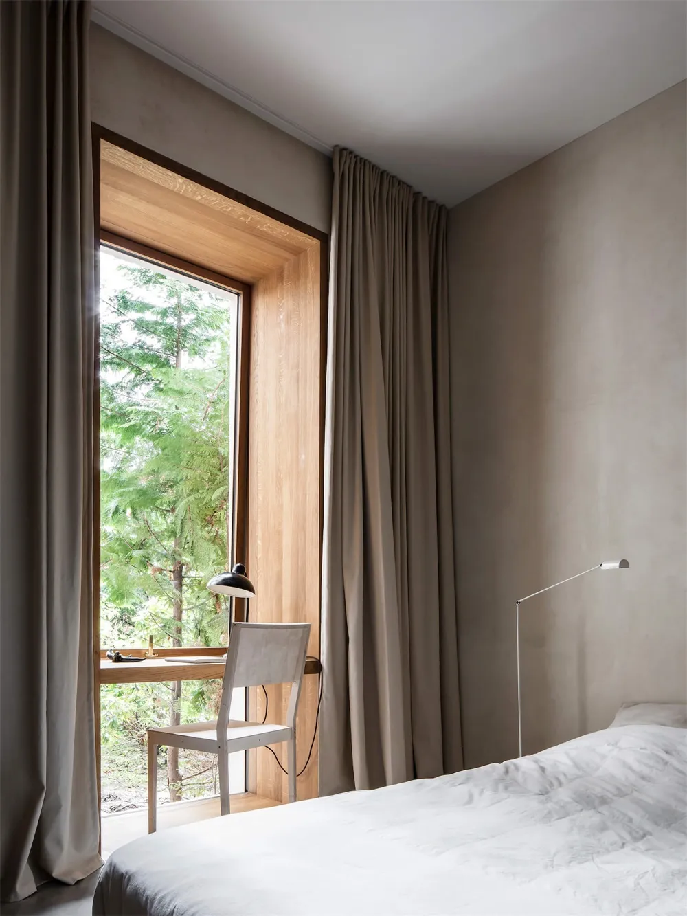 A minimalist bedroom with neutral-colored walls, curtains, and bedding. A small desk with a lamp and chair is placed next to