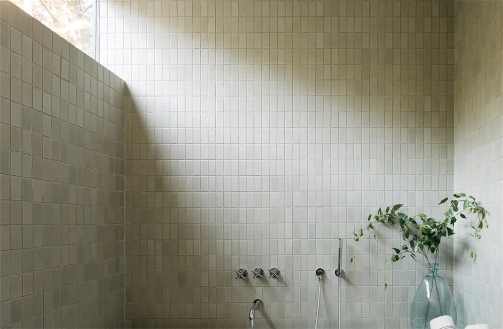 A modern bathroom with white tiled walls and floor, featuring a vase with green leaves and a window letting in natural light.