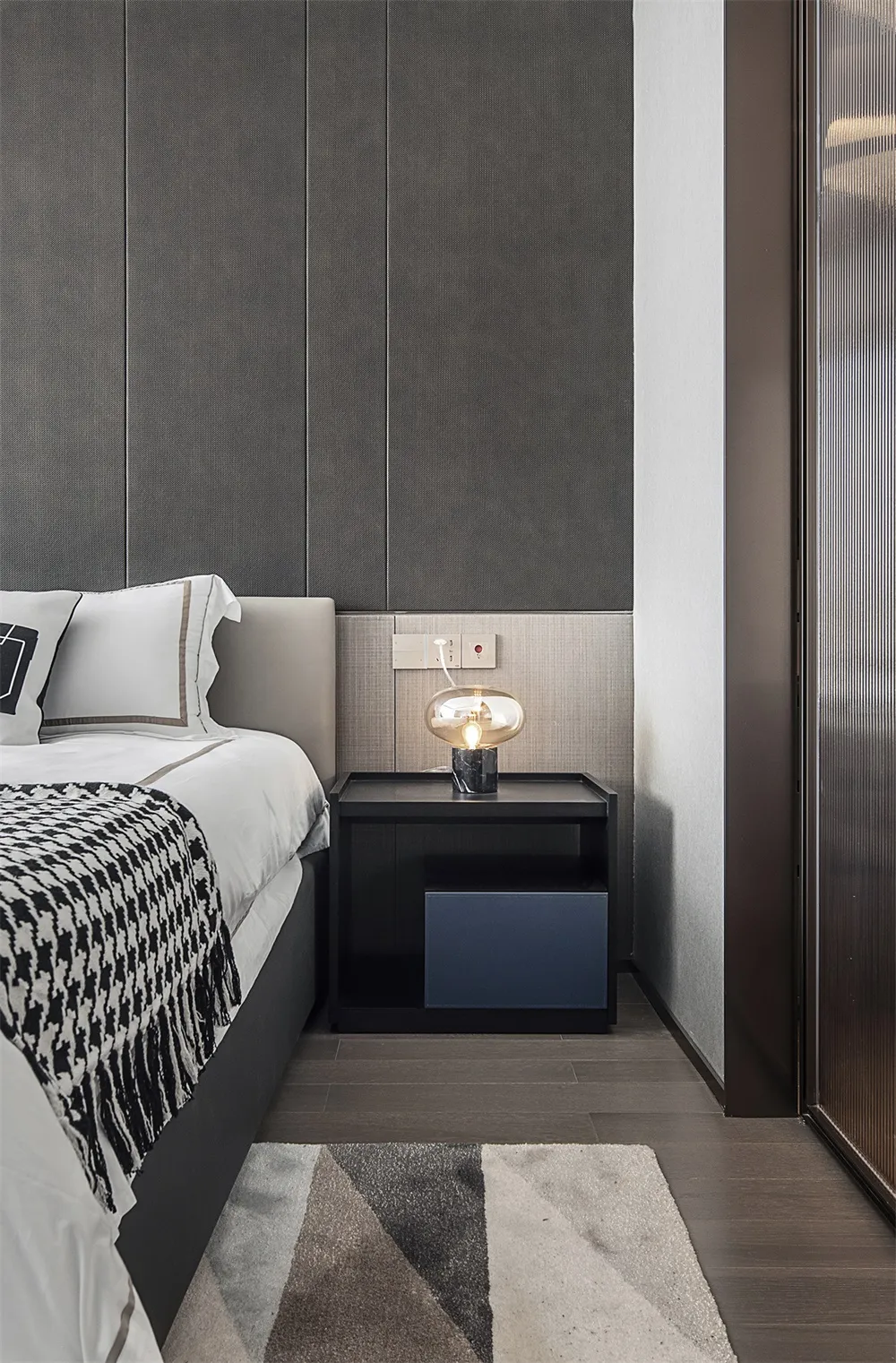 A minimalist bedroom with a bed, nightstand, and lamp, featuring gray fabric, glass, and metal materials.