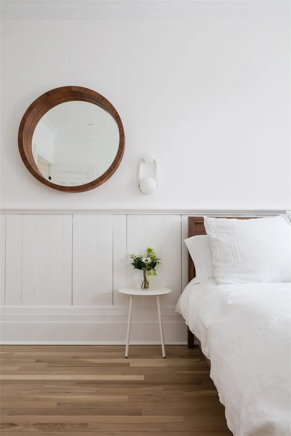 A minimalist bedroom featuring a bed with white bedding, a wooden headboard, and a small white side table with a vase of flow