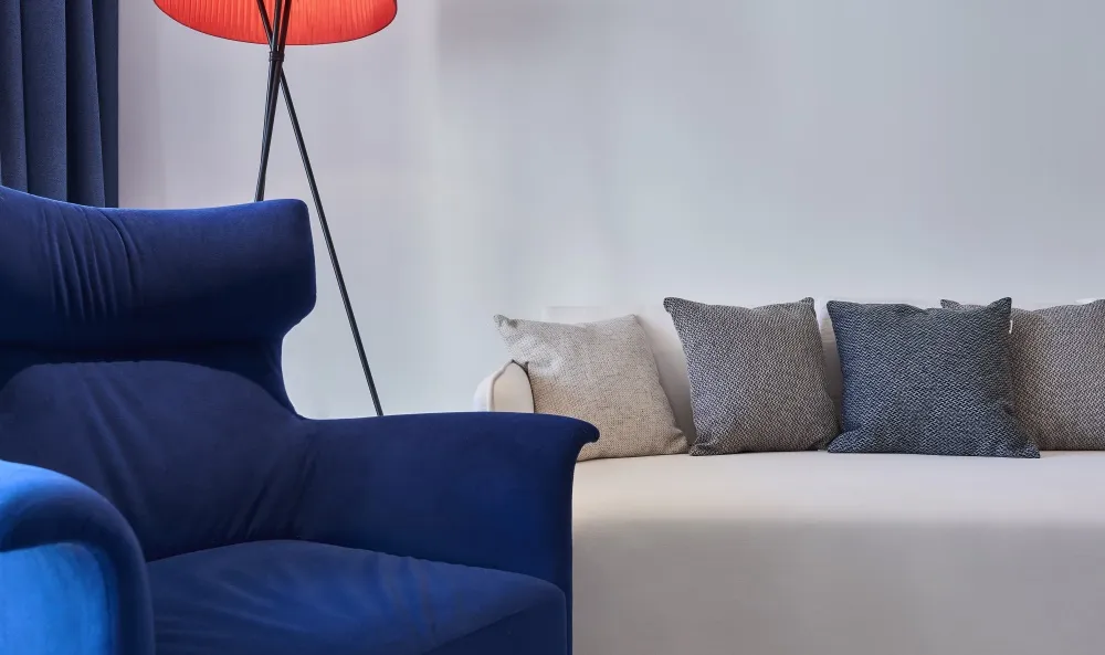 A modern living room with a blue armchair, a beige sofa with three cushions, and a red floor lamp.