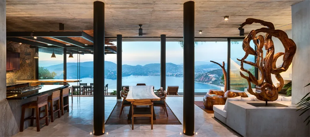 A modern residential dining room featuring a concrete floor and ceiling, with large windows overlooking a scenic landscape.