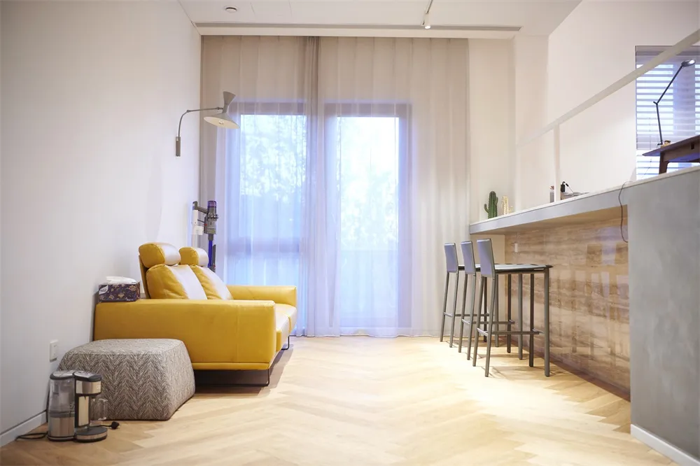 A spacious, modern living room with a yellow sofa, hardwood floors, and sheer curtains.