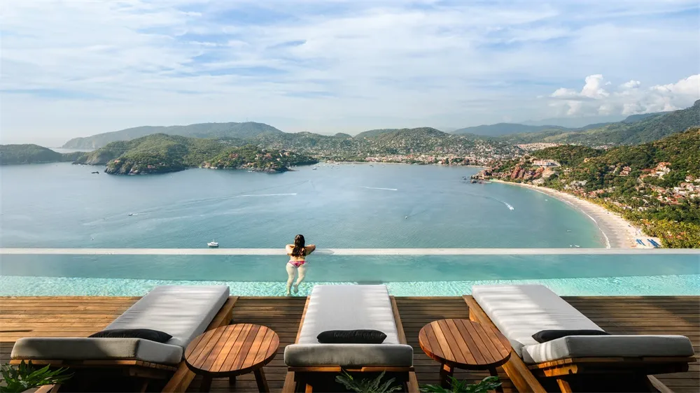 A woman in a swimsuit standing on the edge of a cliffside pool overlooking the ocean and mountains.
