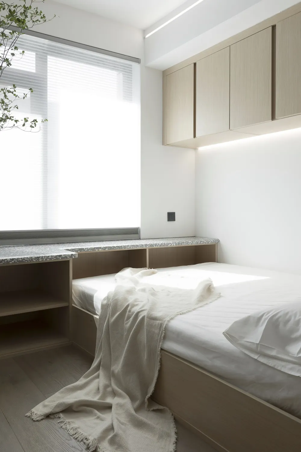 A minimalist white bedroom with a wooden bed, white bedding, and a light-colored cabinet above it, featuring a large window w