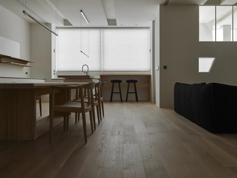 A modern minimalist living room with wooden flooring, featuring a black sofa and a kitchen area with a white countertop and s
