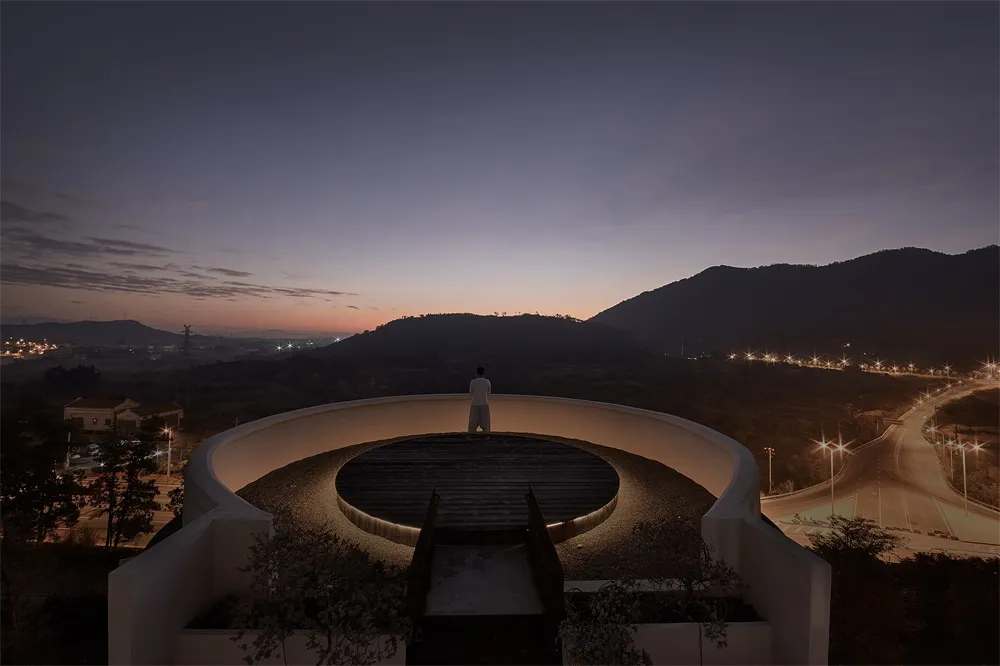 A person standing on a circular terrace overlooking a valley at sunset.