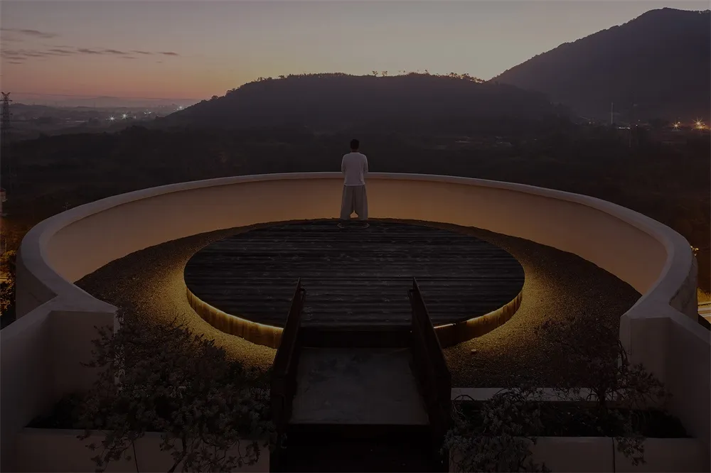 Person standing on a circular wooden platform with illuminated edges, overlooking a mountainous landscape at sunset.