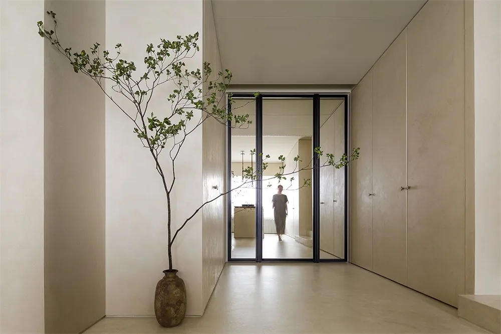 A minimalist hallway with natural light filtering through a window and a tall, slender tree vase with green leaves adding a t