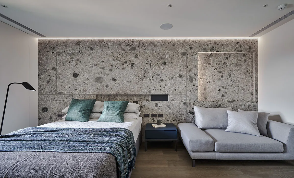 A modern bedroom featuring a concrete wall, a cozy bed with blue and grey bedding, and a comfortable grey couch.