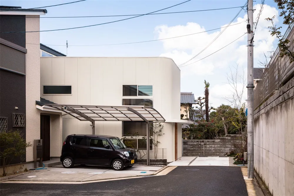 A black car parked under a metallic carport in front of a two-story modern house with a flat roof and large windows.