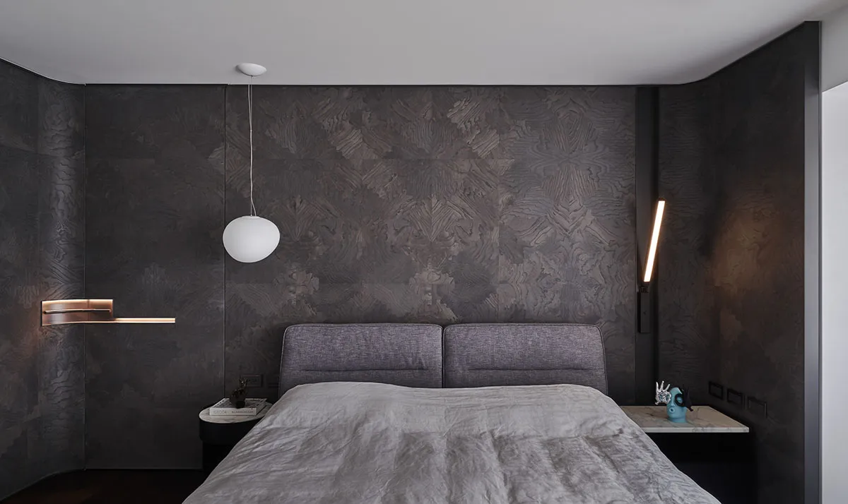A modern bedroom featuring a textured dark wall, a grey upholstered bed, and minimalistic design elements.